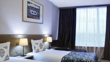 Habitación de hotel con dos camas, cabecera, lámparas de noche, un ventanal con cortinas y un cuadro en la pared.