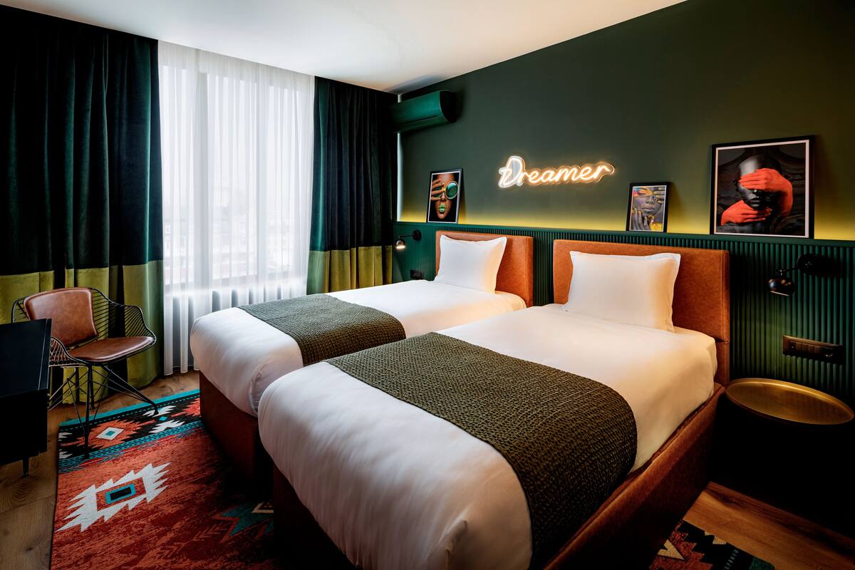 A hotel room with two beds, a chair, a neon sign, and artwork on the wall.