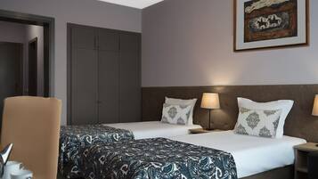 Une chambre d’hôtel avec deux lits, un coin salon avec un fauteuil, une table avec une tasse et une soucoupe, et un tableau accroché au mur.