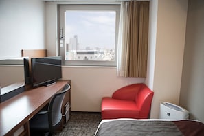 A hotel room with a desk, a television, a red chair, a bed, and a window with a city view.