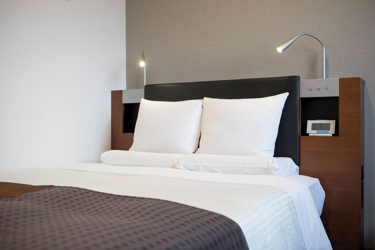 Ein ordentlich bezogenes Bett mit weißen Laken, einem dunklen Kopfteil und einem Nachttisch mit Lampe und Uhr.