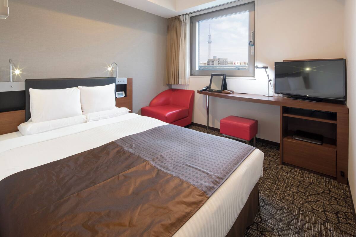 Ein Hotelzimmer mit einem Bett, einem roten Sessel, einem Schreibtisch mit Fernseher und einem Fenster mit Blick auf die Stadt.