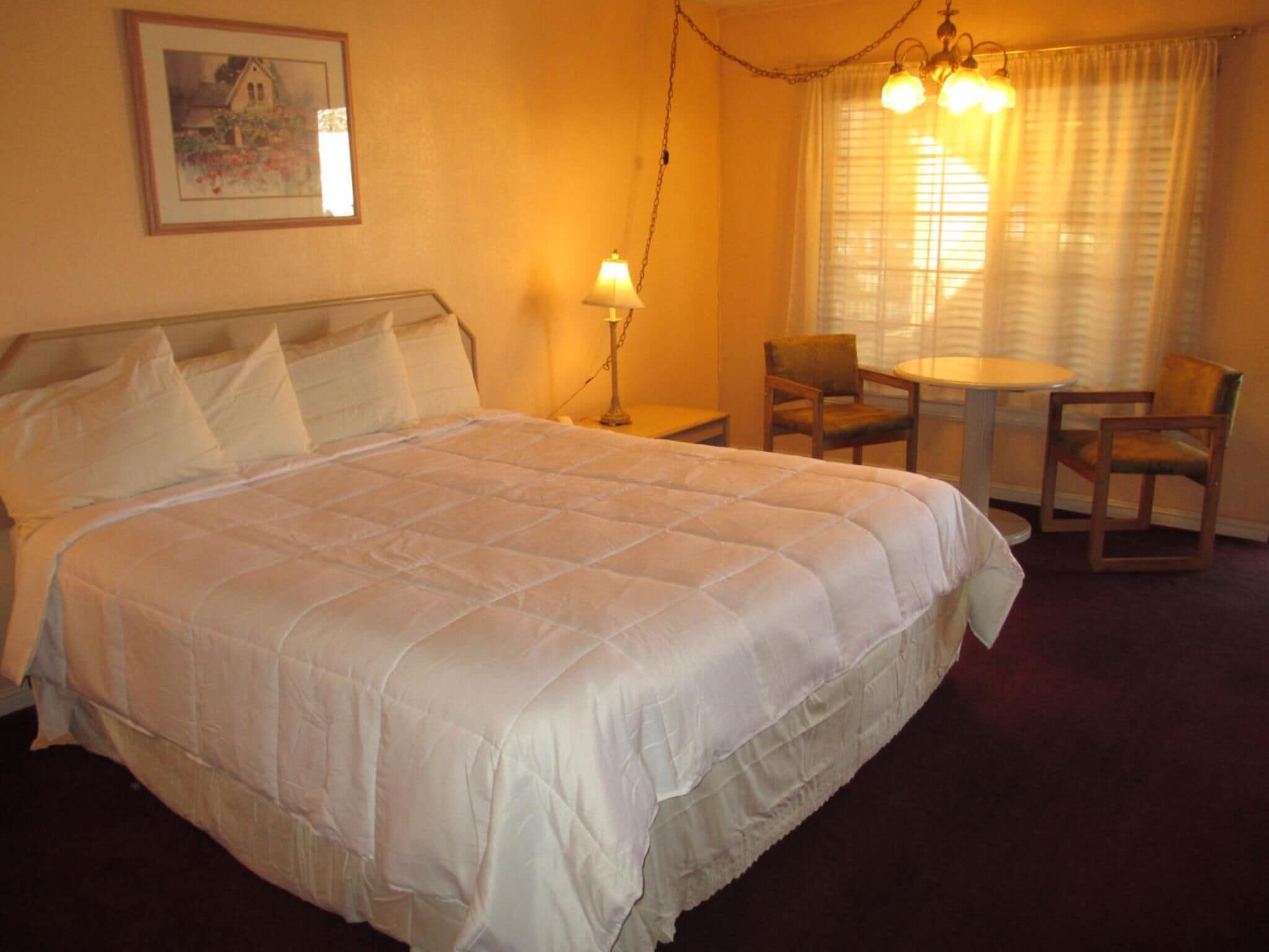 A hotel room with a large bed, a small table, two chairs, a lamp, and a painting on the wall.