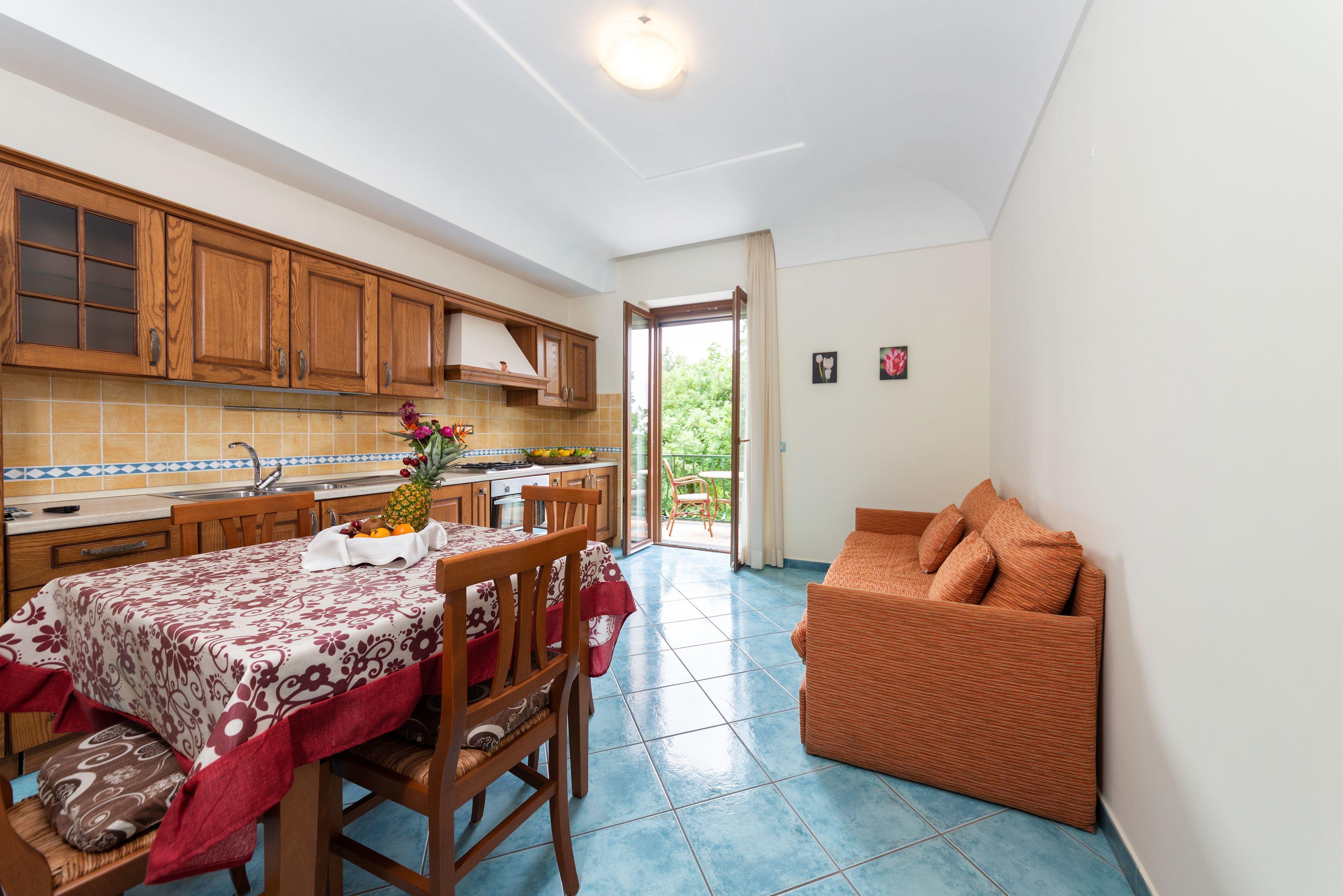 A kitchen with wooden cabinets, a dining table with a floral tablecloth, and a sofa in a living area.