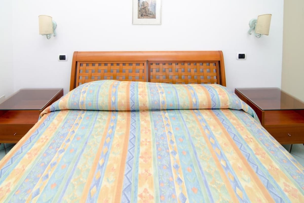 Un llit amb un sobrell amb motius, dues taules de nit i làmpades penjades a la paret.