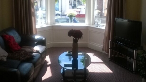 A living room with a sofa, a glass coffee table with a vase, a TV, and a window with signs indicating 'NO VACANCIES'.