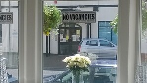 A hotel entrance with a 'NO VACANCIES' sign, a car parked outside, and a vase of flowers on the windowsill.