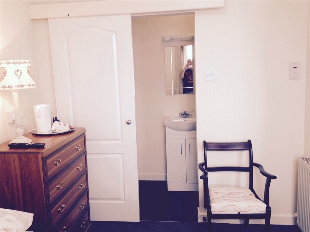 A bedroom with a wooden dresser, a chair, a bedside table with a lamp, and a door leading to a bathroom with a sink and mirror.