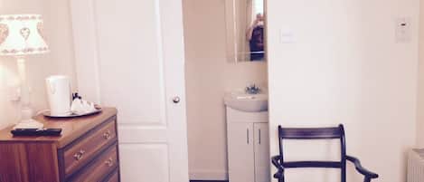 A bedroom with a wooden dresser, a chair, a bedside table with a lamp, and a door leading to a bathroom with a sink and mirror.