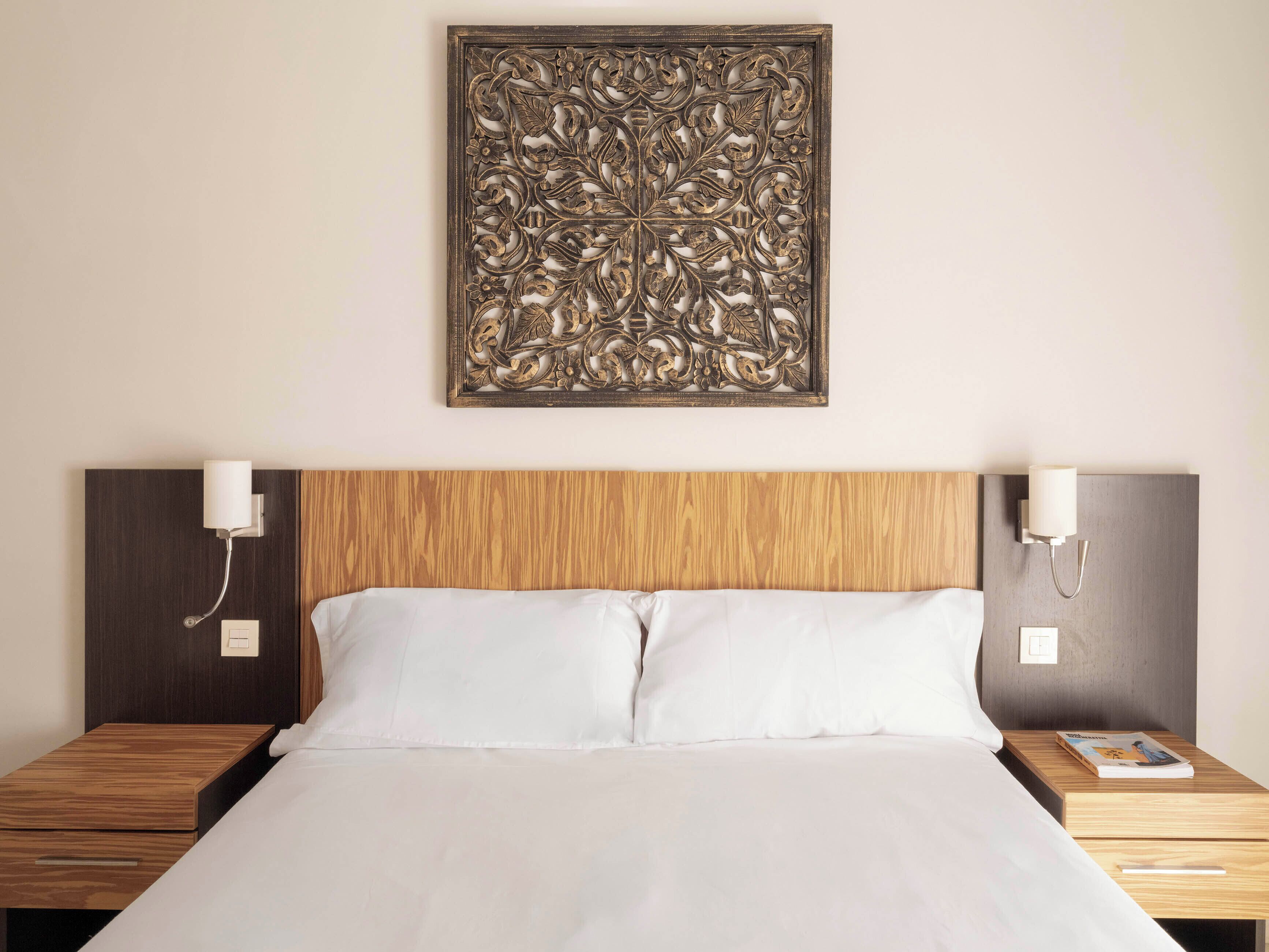 A hotel room with a wooden headboard, two bedside tables, a large bed with white linens, and a decorative wall piece above the bed.