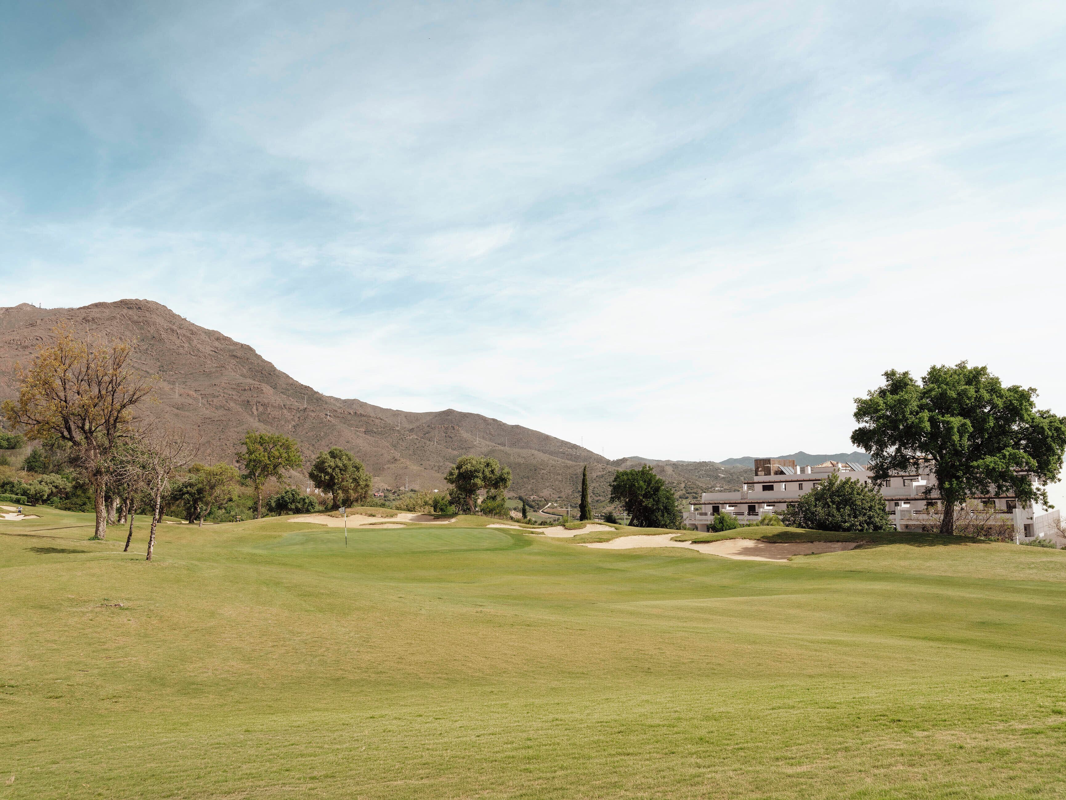 Un campo de golf con montañas al fondo, un edificio de varios pisos y un cielo despejado.