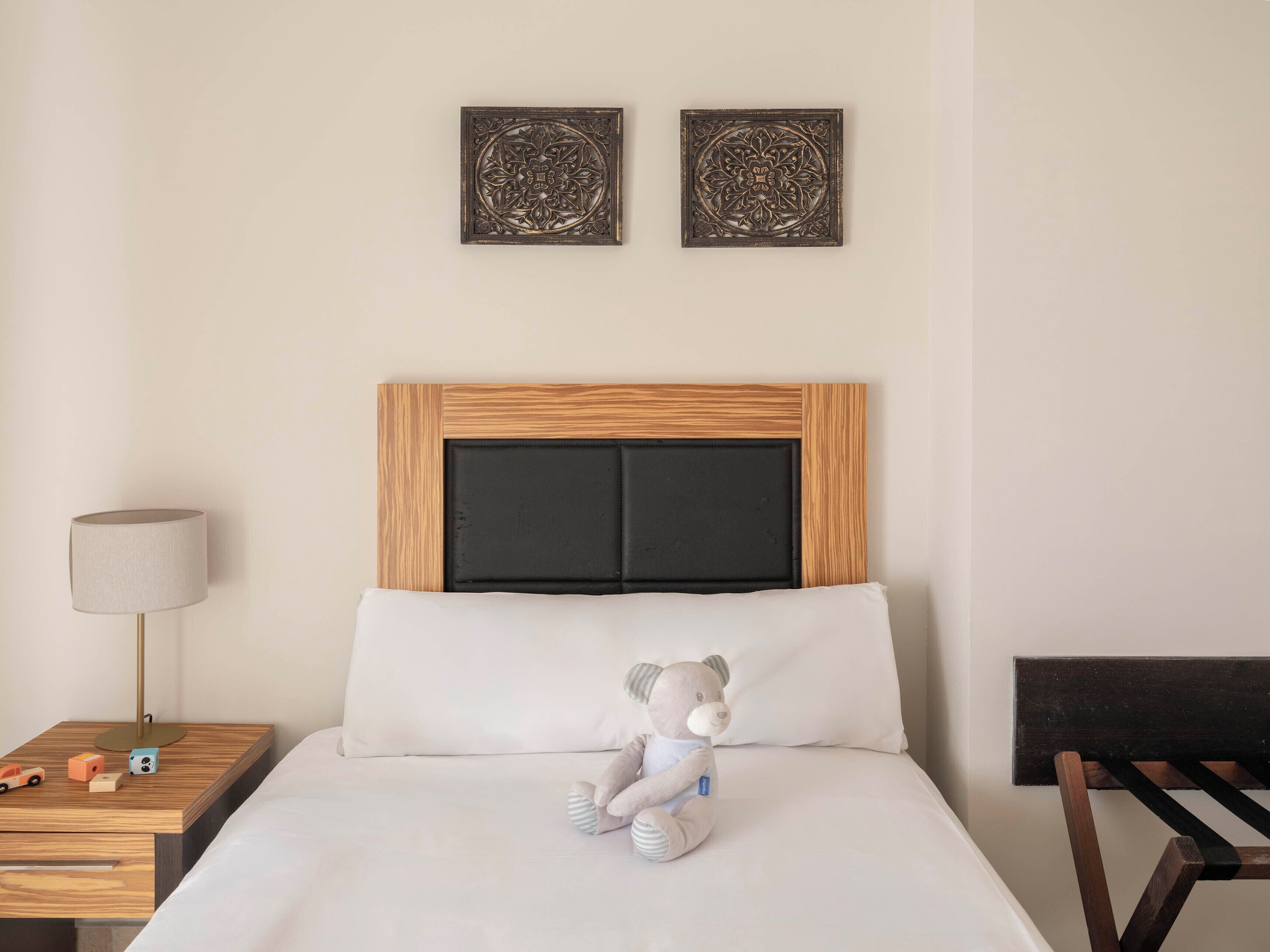 Una habitación de hotel con cama, mesita de noche, lámpara, un osito de peluche y dos adornos en la pared.