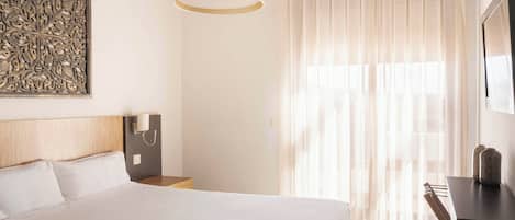 A hotel room with a large bed, a bedside table, a lamp, a decorative wall piece, and a window with sheer curtains.