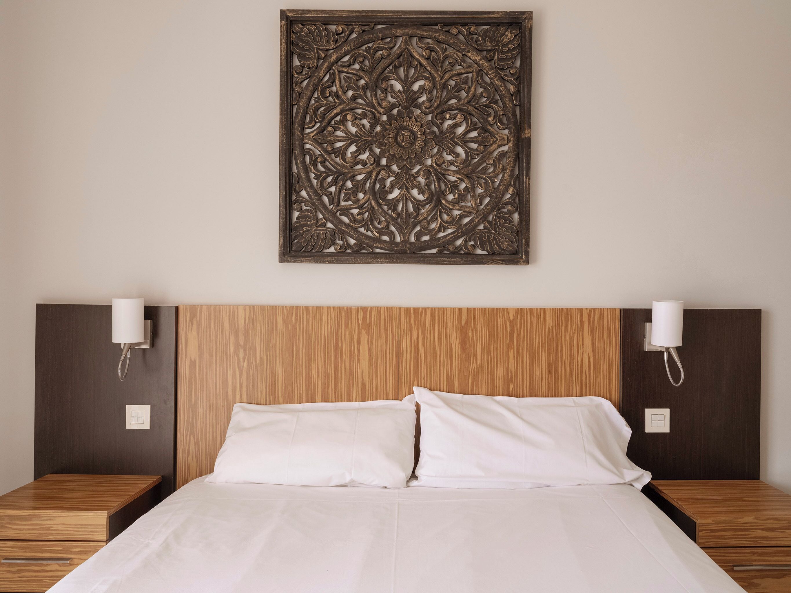 A hotel room with a wooden headboard, two bedside tables with lamps, and a decorative wall piece above the bed.