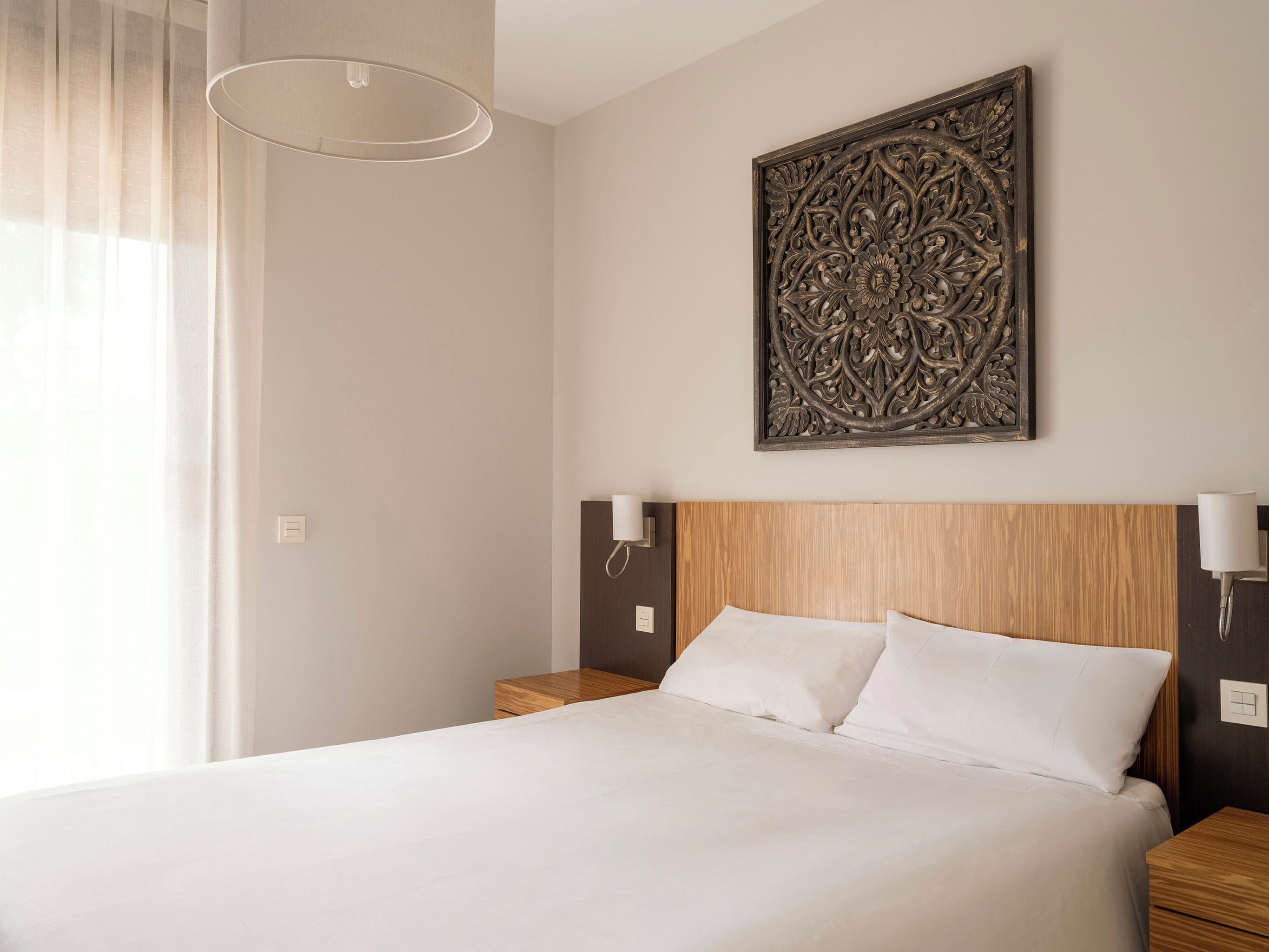 A hotel room with a bed, bedside lamps, a decorative wall piece, and a window with curtains.