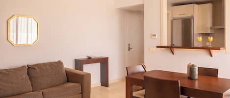 A modern living room with a brown sofa, a dining table with chairs, a small side table, and a kitchen area visible through an open door.