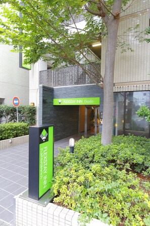 Entrada de un hotel con un cartel de FlexStay Inn Ekoda, rodeada de vegetación y una fachada de edificio con tejas.