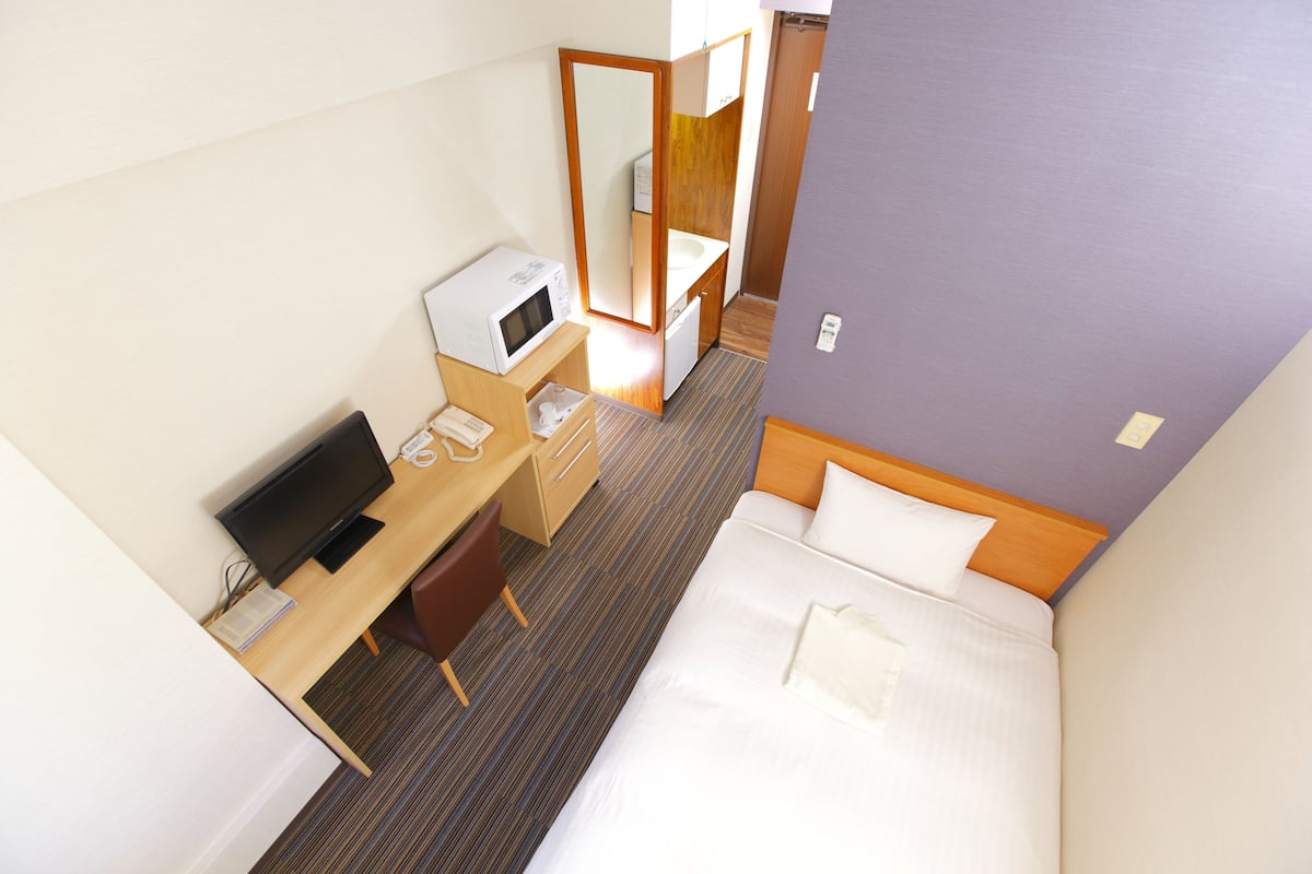 A hotel room with a bed, a desk with a computer, a microwave, a small sink, and a mirror.