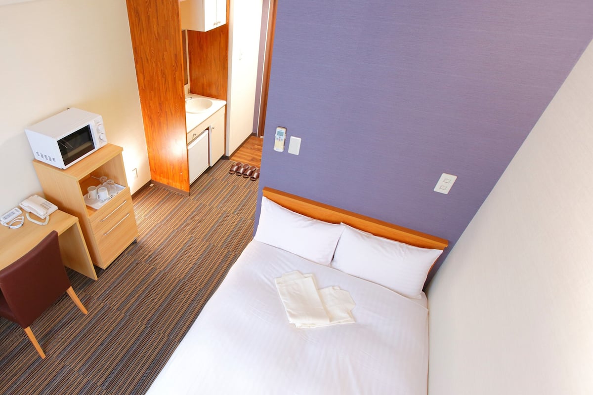 A hotel room with a bed, a desk with a microwave, a vanity with a mirror, and a shoe rack.