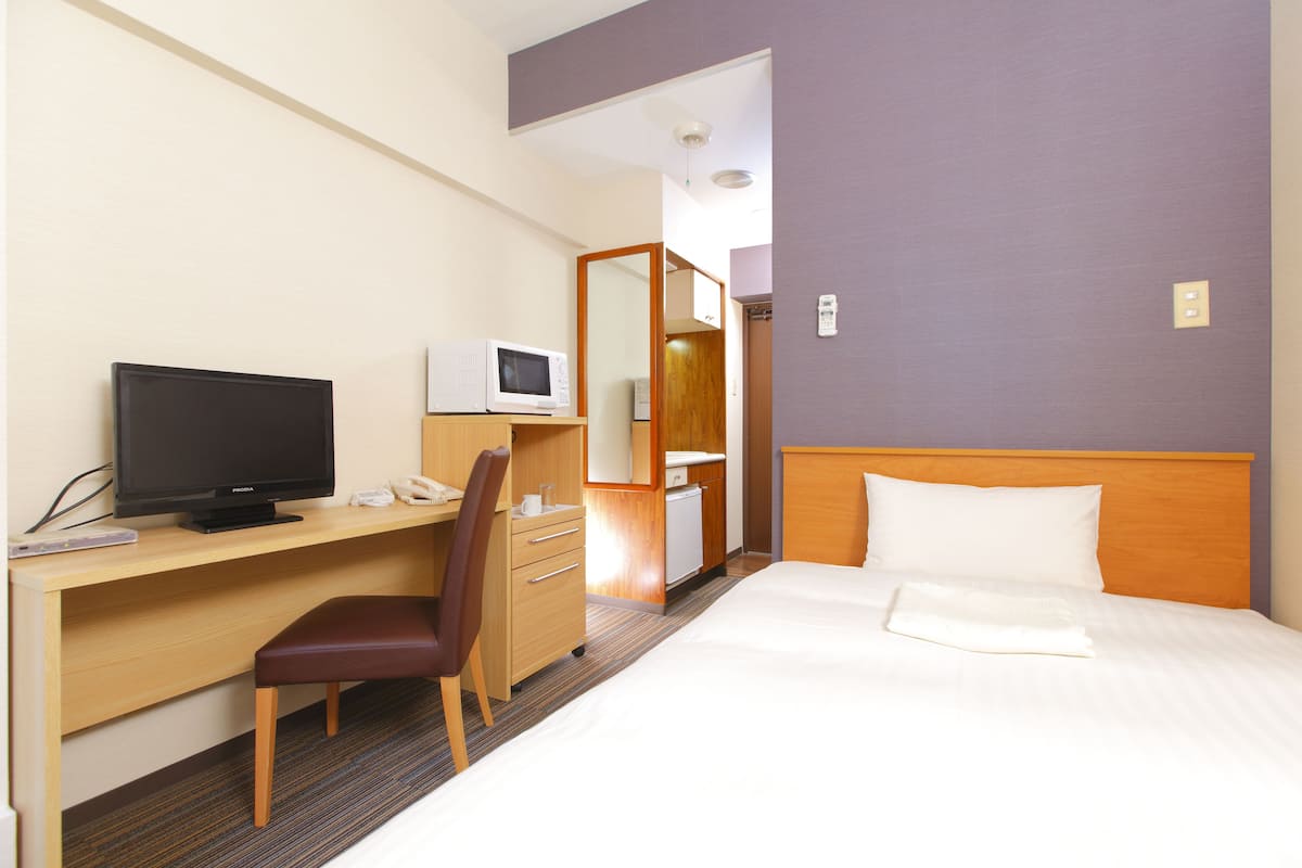 A hotel room with a bed, a desk with a television, a chair, a microwave, and a small bathroom.