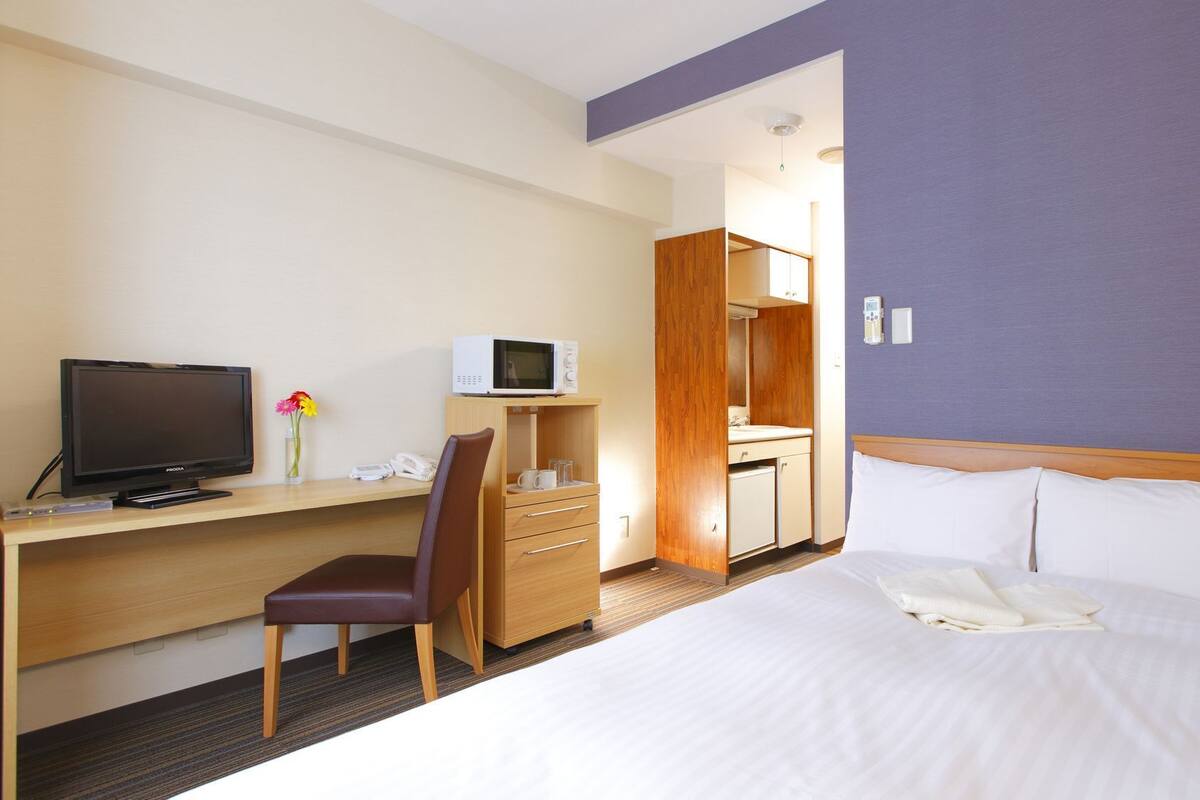 A hotel room with a bed, a desk with a television, a chair, a microwave, a mirror, and a bathroom.