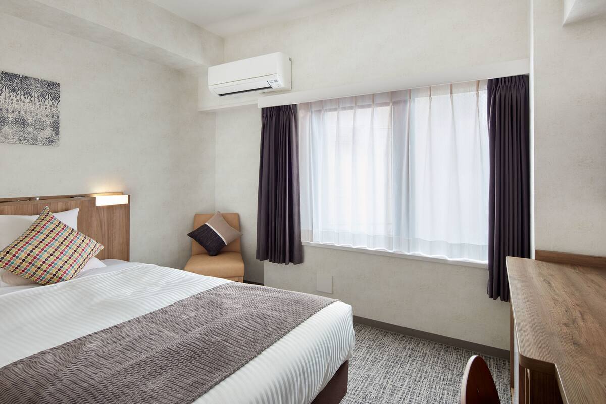 Una habitación de hotel con una cama, un escritorio, aire acondicionado y una ventana con cortinas.