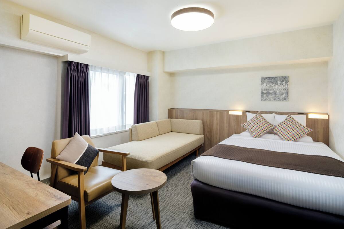 Una habitación de hotel con cama, sofá, sillón, mesita y ventana con cortinas.