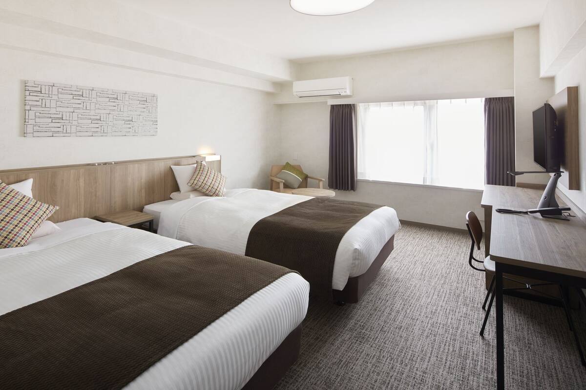 Habitación de hotel con dos camas, un escritorio con silla, televisión y aire acondicionado.