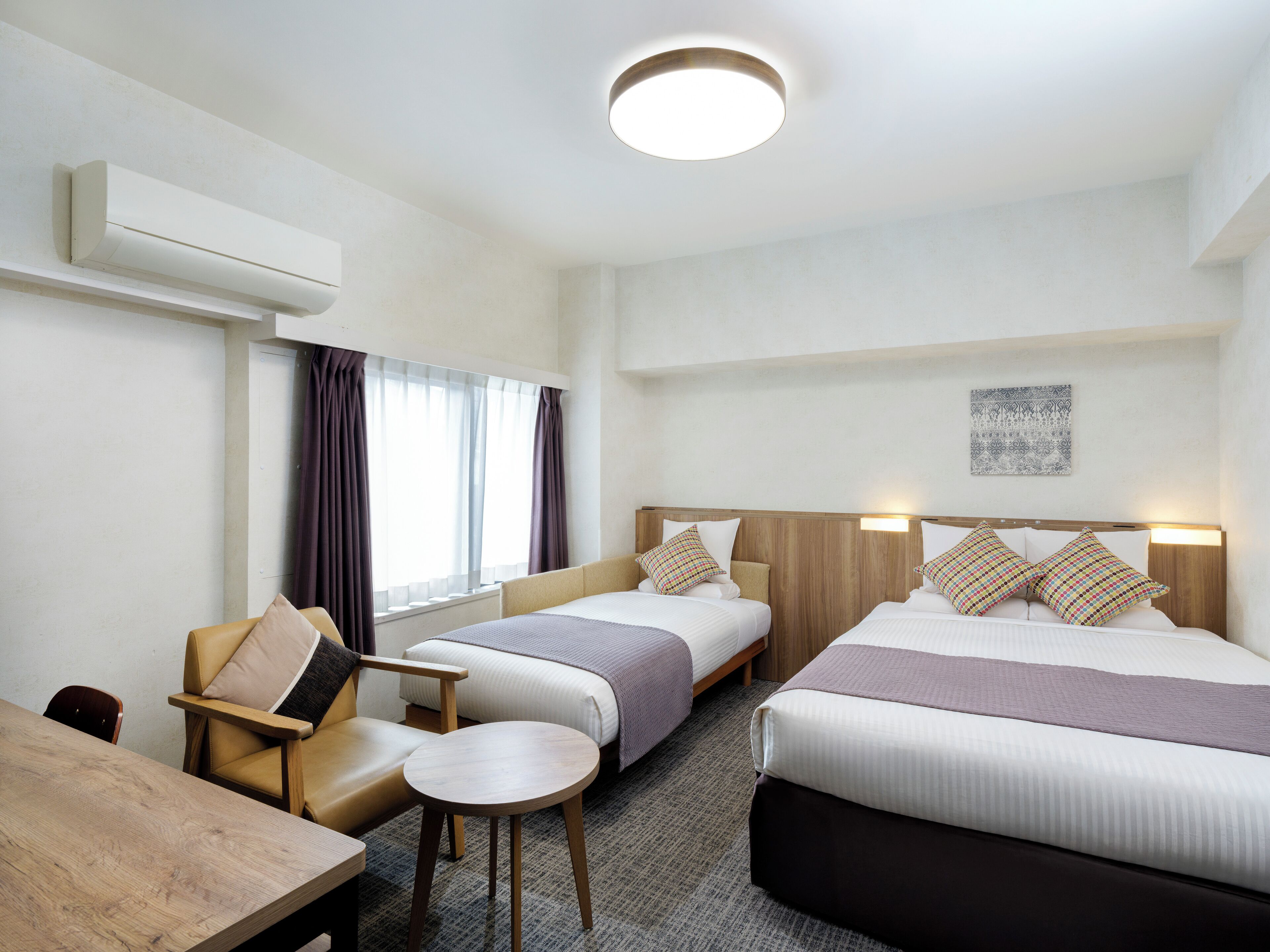 A hotel room with two beds, a small table, a chair, and a window with curtains.
