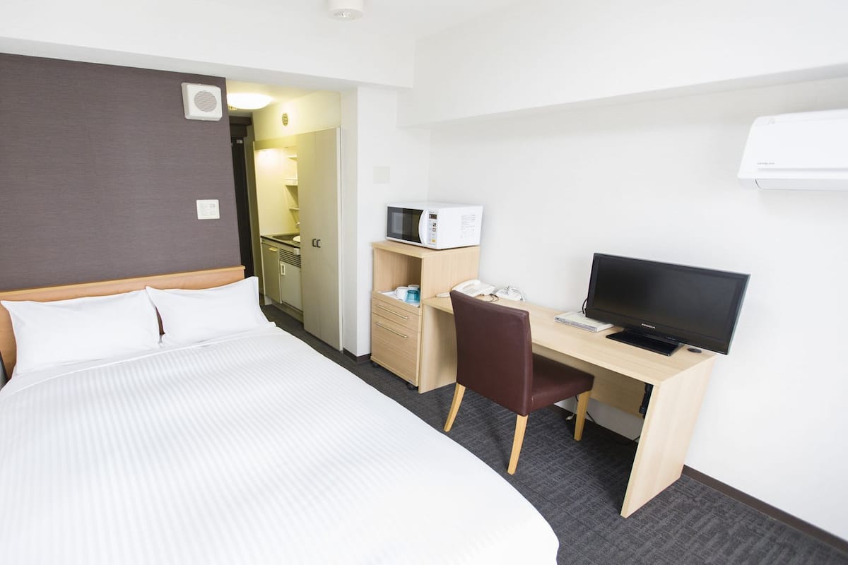 A hotel room with a bed, a desk with a computer and a television, a microwave, and a small kitchenette.