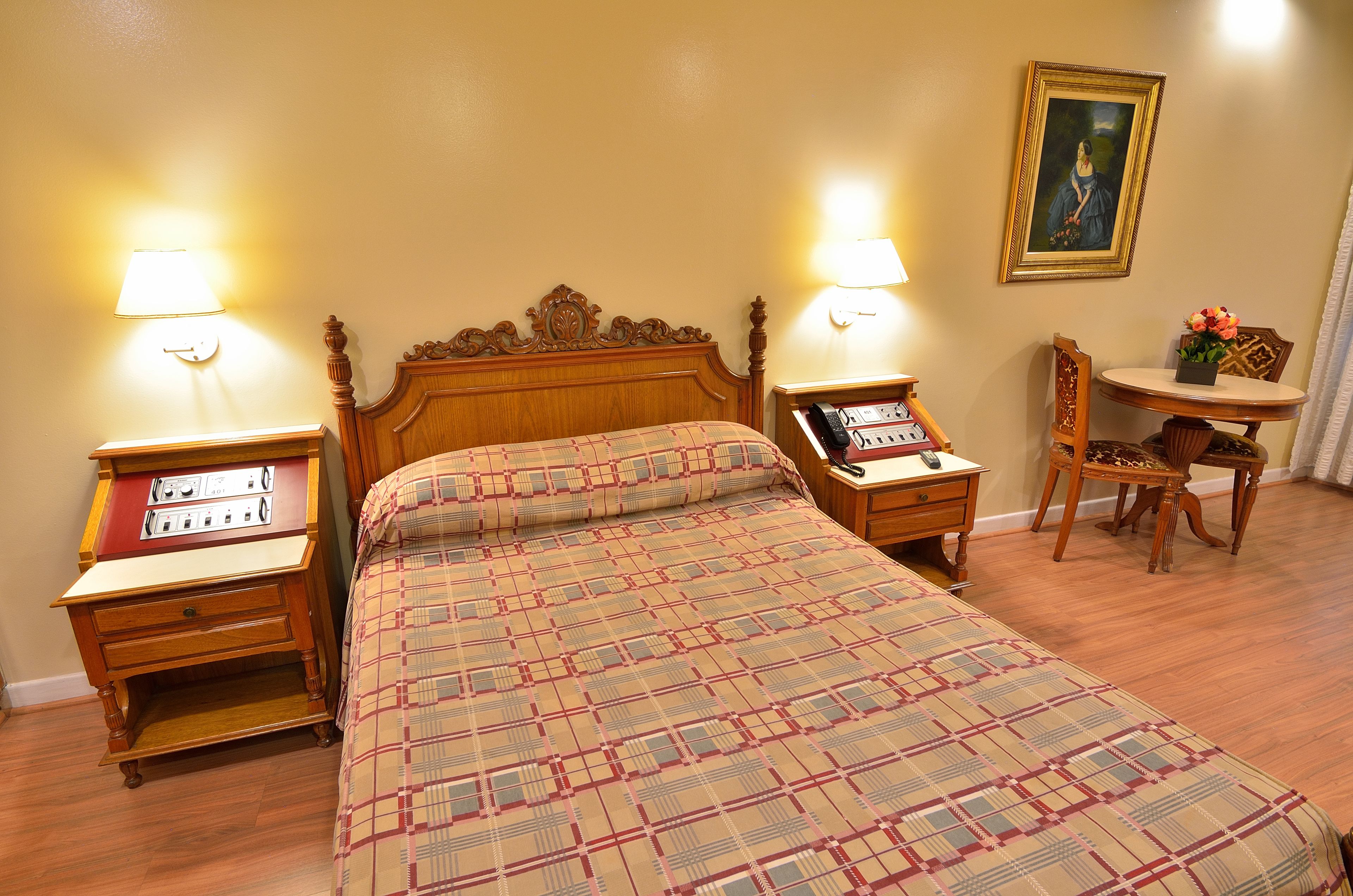 A hotel room with a wooden bed, two bedside tables with lamps, a chair, a small table with a vase of flowers, and a framed picture on the wall.