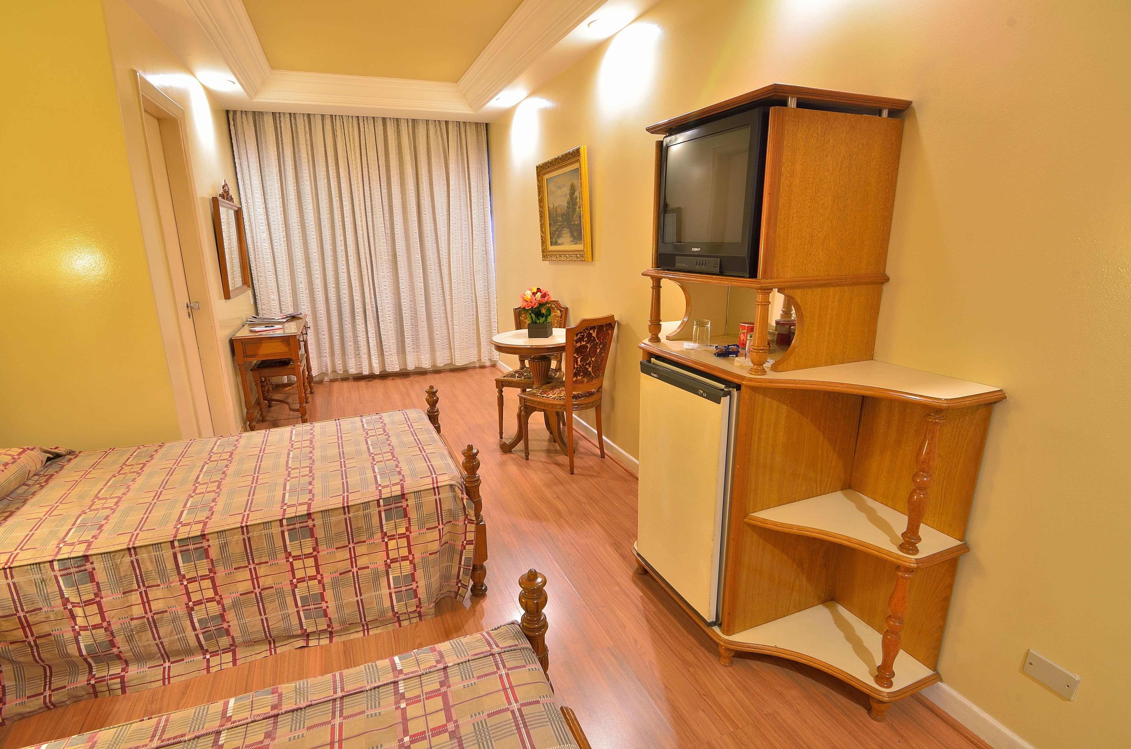 Ein Hotelzimmer mit zwei Betten, einem Holzkoffer, einem Fernseher und einem kleinen Tisch mit Stuhl.