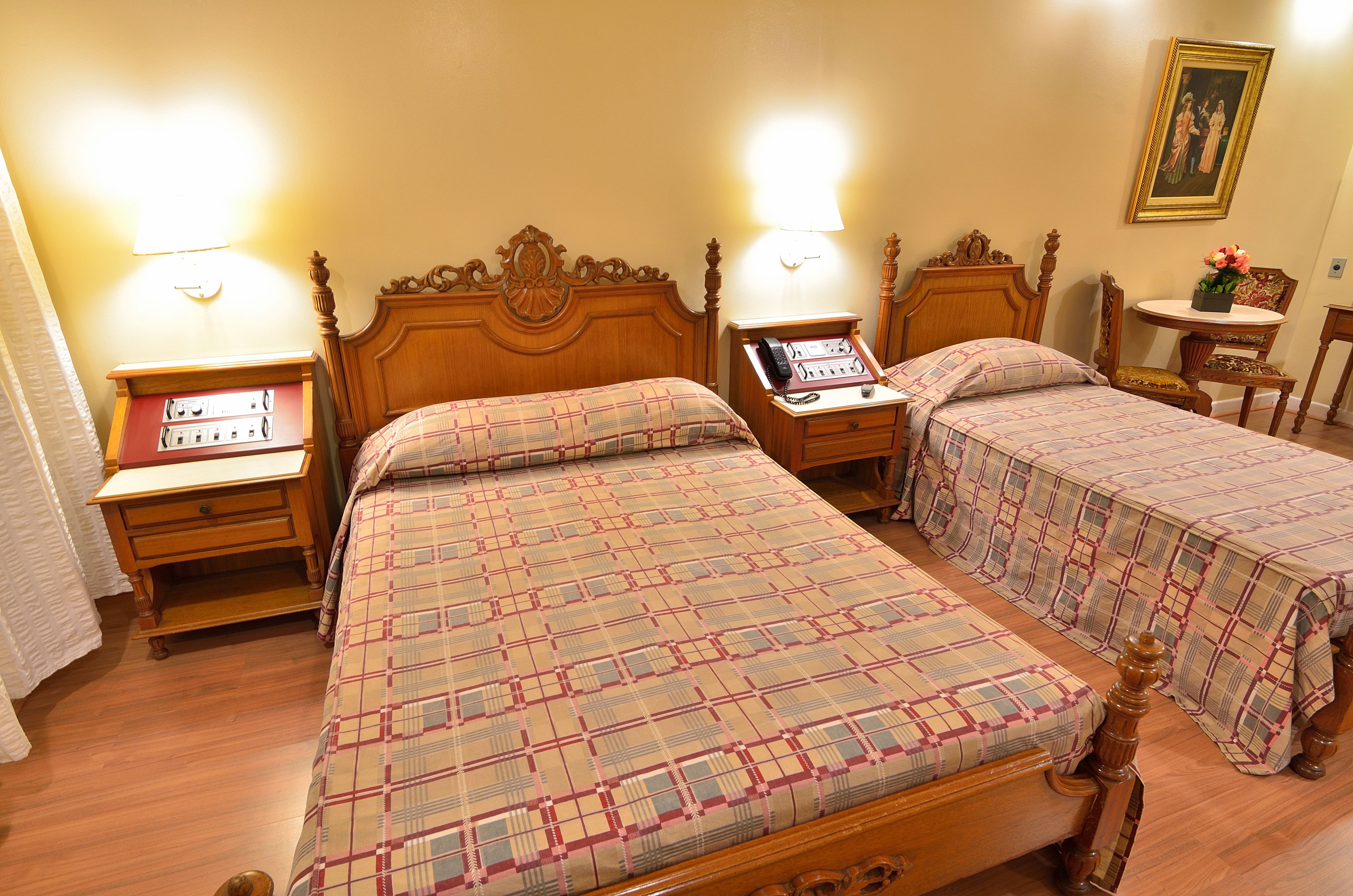 A hotel room with two wooden beds with matching headboards, bedside tables with lamps, a small round table with a flower vase, and a framed picture on the wall.