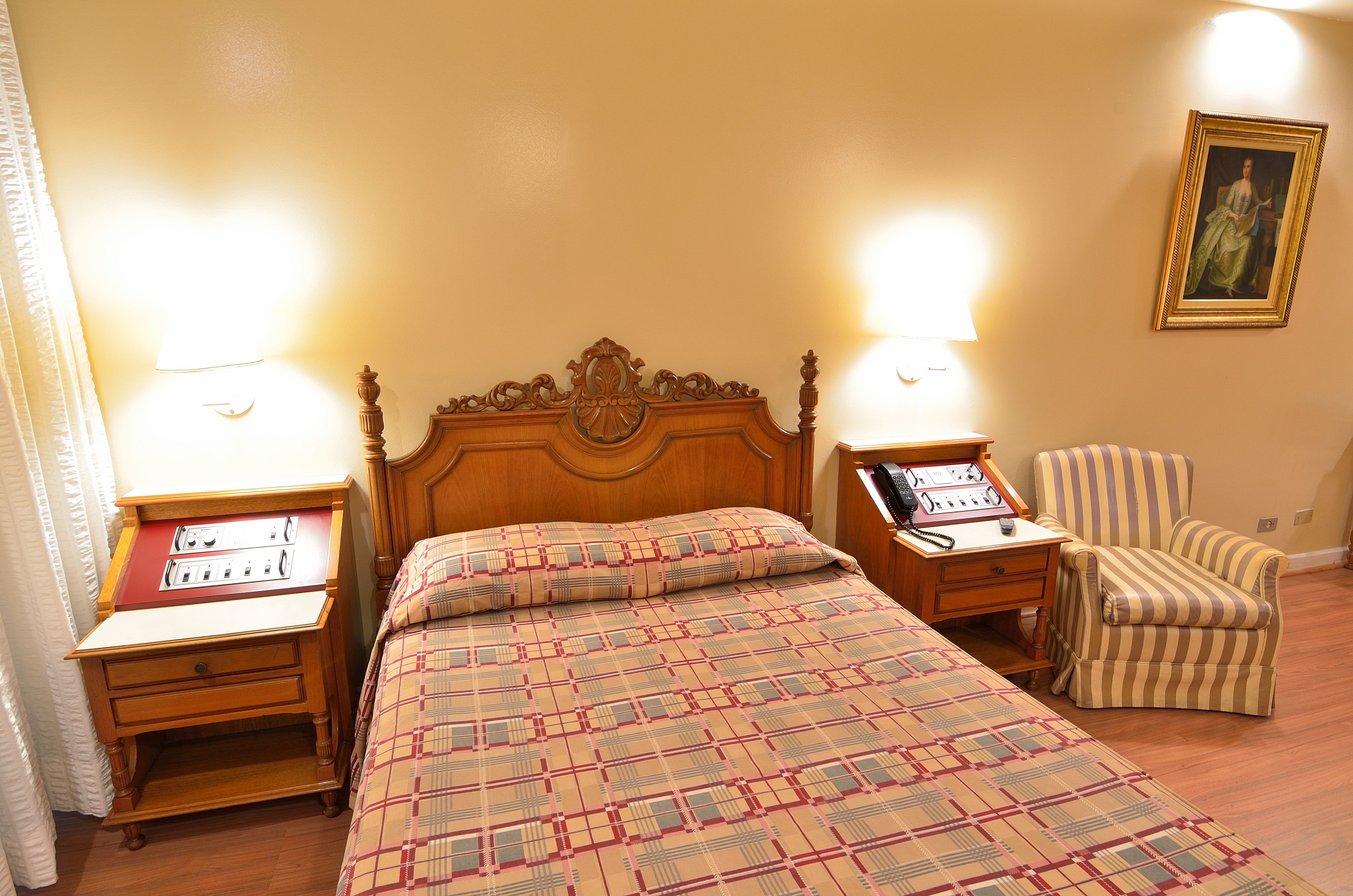 A hotel room with a wooden bed, two bedside tables with lamps, a striped armchair, and a painting on the wall.