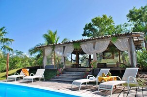 Un área junto a la piscina con sillones de descanso, una zona de estar cubierta y una piscina.