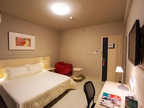 Quarto de hotel com cama, escrivaninha com cadeira, poltrona vermelha, mesa redonda pequena, TV fixada na parede e quadro pendurado na parede.