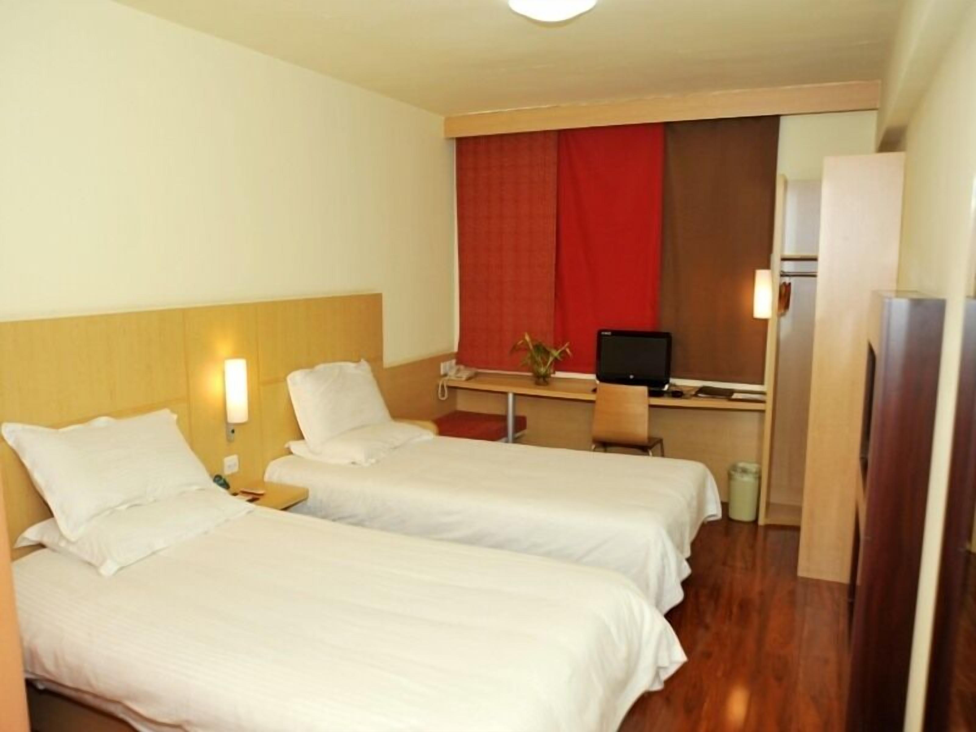 A hotel room with two single beds, a desk with a computer, a mirror, and a refrigerator.