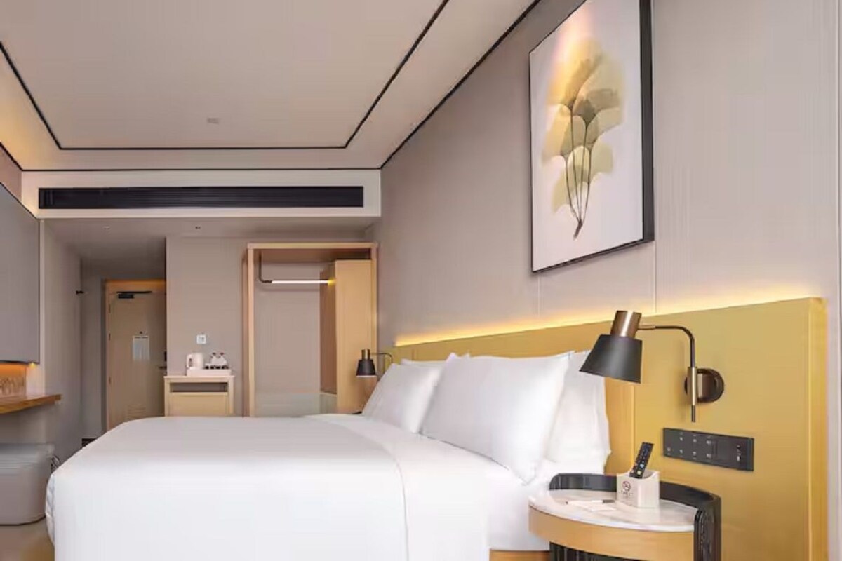 Ein modernes Hotelzimmer mit einem großen Bett, einem Nachttisch mit Lampe, einem kleinen Schreibtisch mit Computer und einem gerahmten Kunstwerk an der Wand.