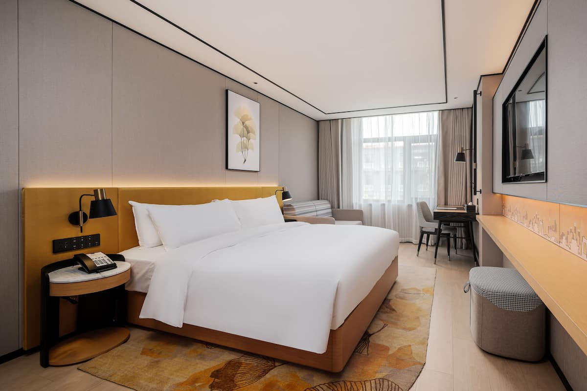 A modern hotel room with a large bed, bedside table with a phone, a desk with a chair, and a large window with curtains.