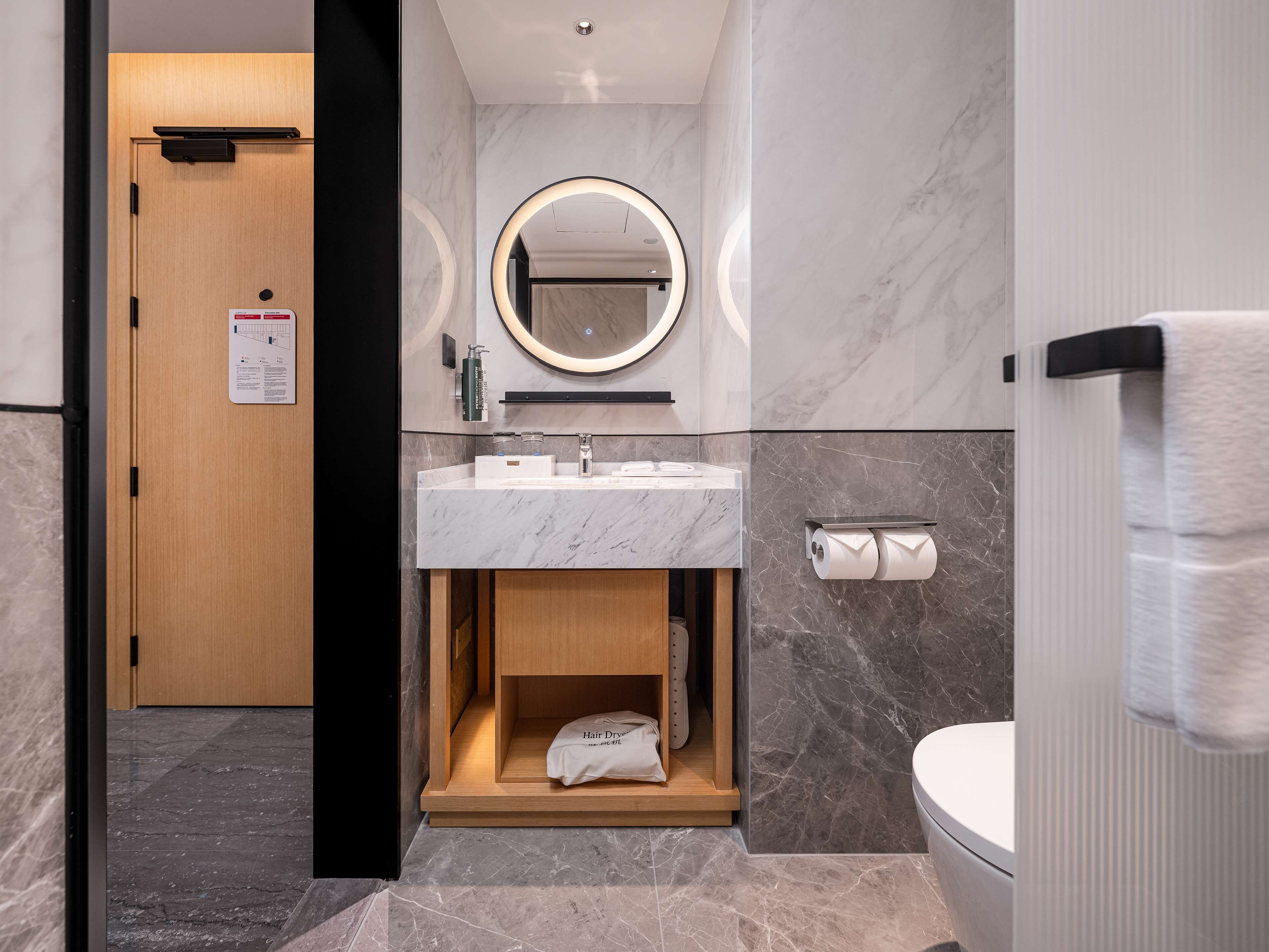 Un baño moderno con un lavabo de mármol, un espejo redondo con iluminación, un inodoro con toallero y un armario de madera con una toalla y un secador de pelo.