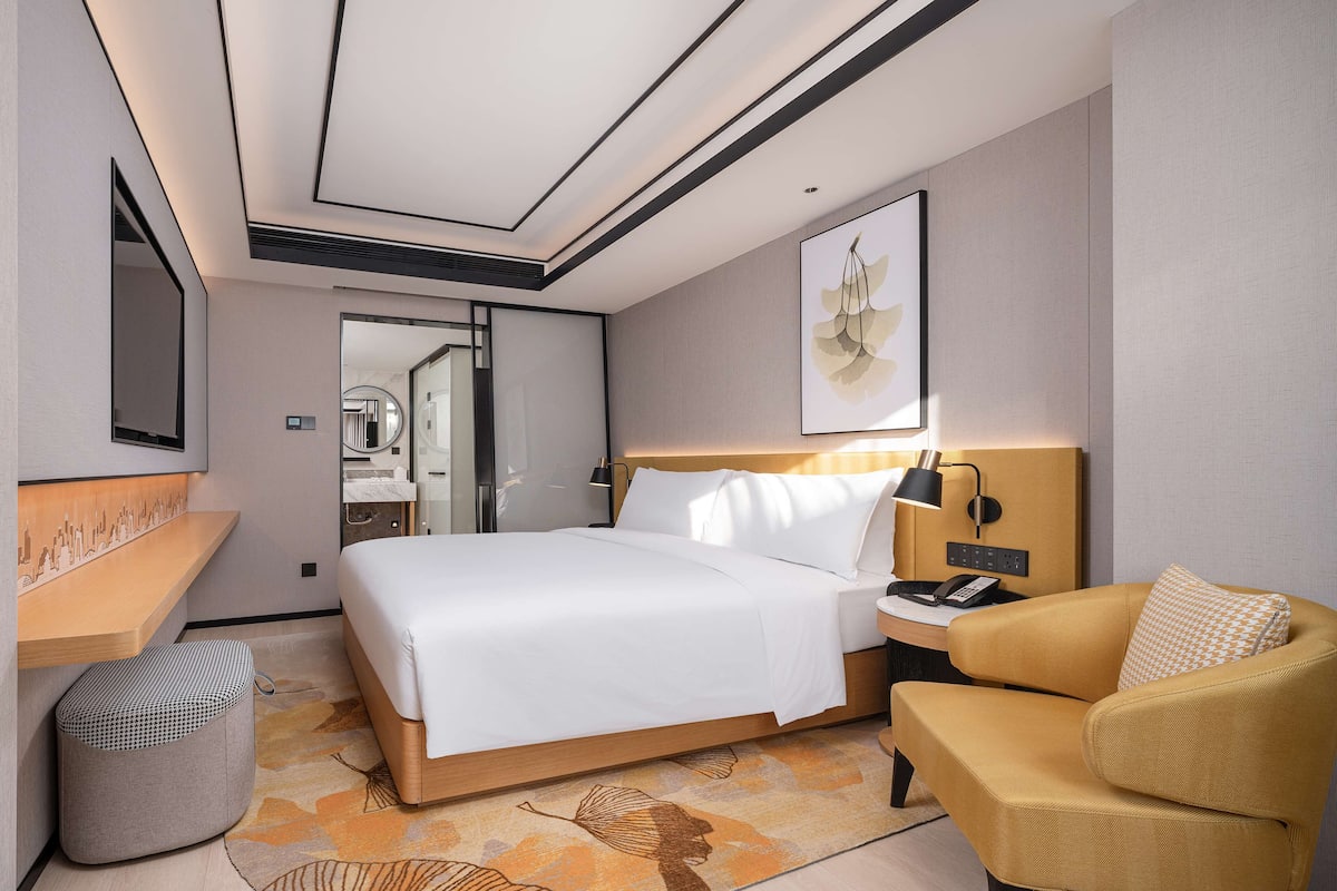 A modern hotel room with a bed, bedside table, lamp, chair, and a view of the bathroom.