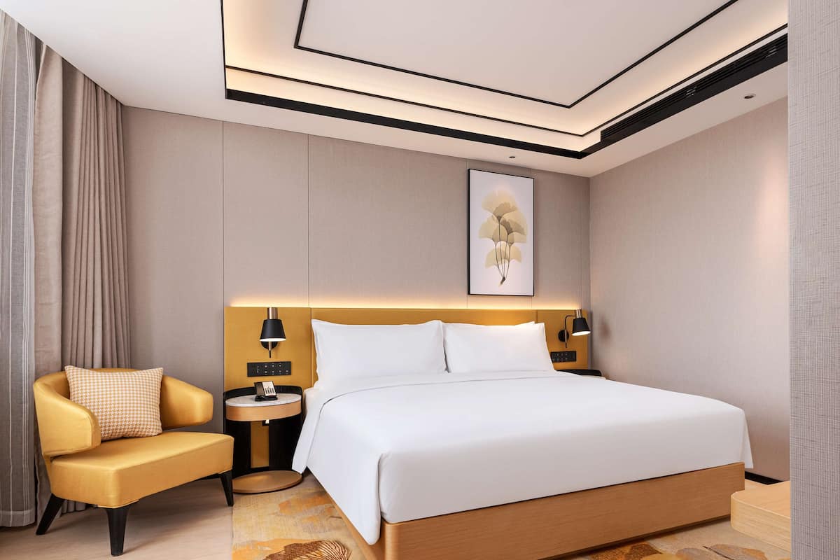 A modern hotel room with a large bed, a sofa, a bedside table with a lamp, and a painting on the wall.