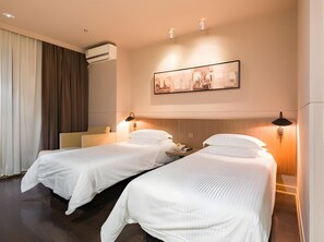 Ein Hotelzimmer mit zwei Betten, einem großen Fenster mit Vorhängen, einer Klimaanlage, einem gerahmten Bild an der Wand und einer Nachttischlampe.