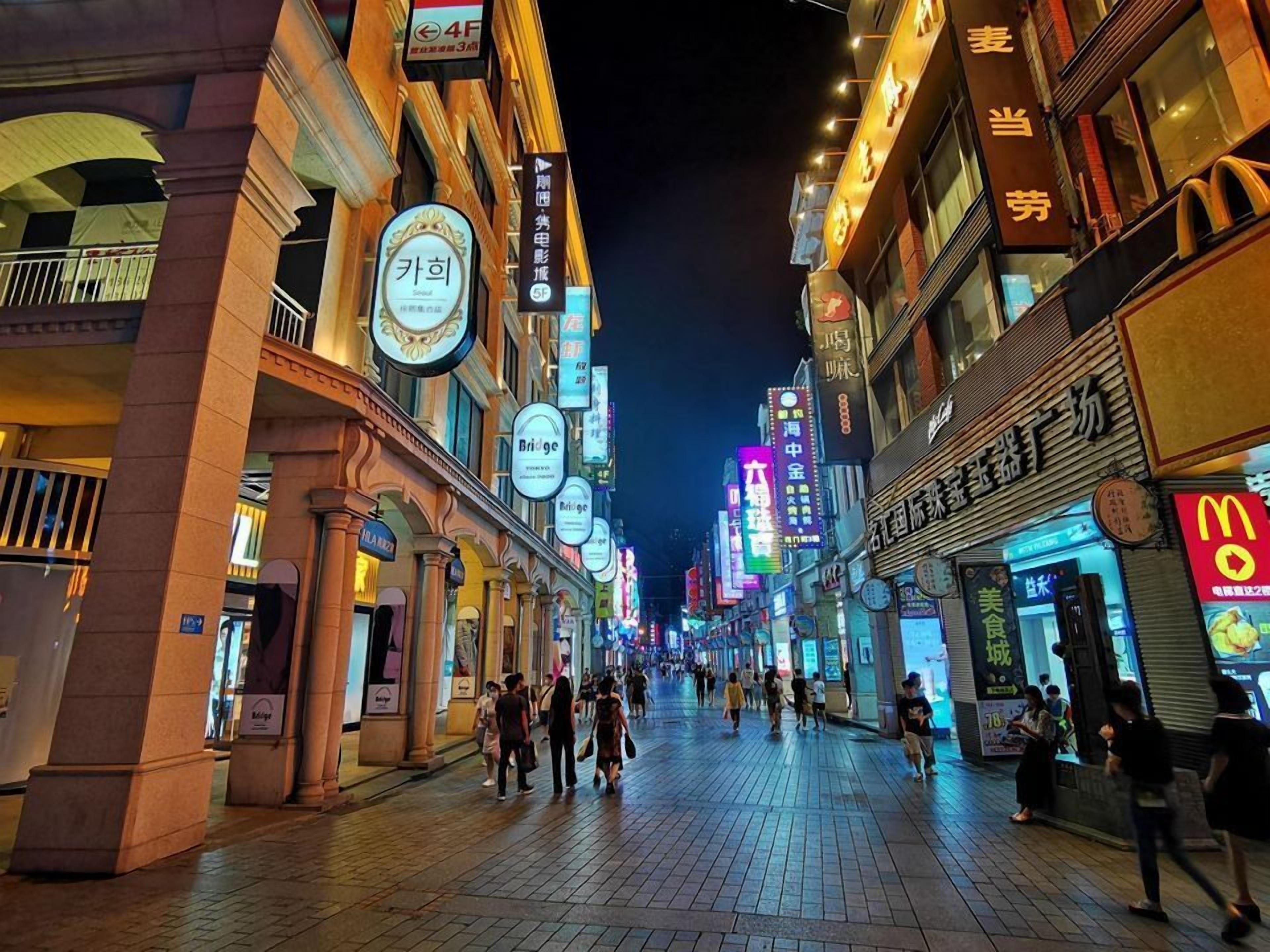 Escena nocturna de una calle con letreros y escaparates iluminados, incluyendo un McDonald's y una señal de puente.