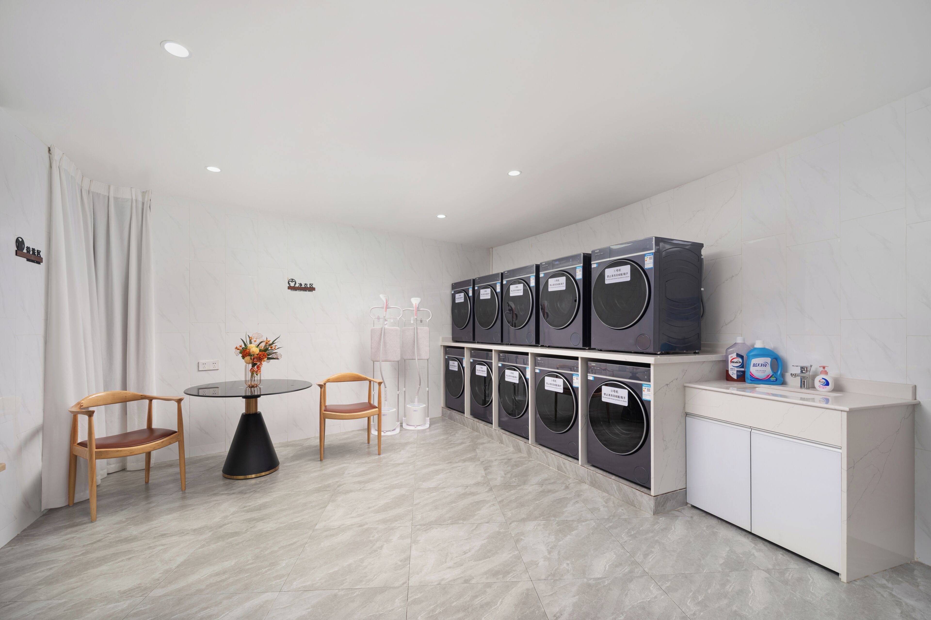 A laundry room with multiple washing machines, a table with chairs, and a cabinet with cleaning supplies.