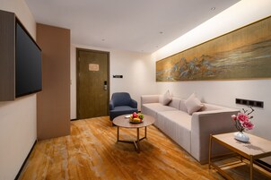 A modern hotel room with a sofa, a round coffee table with fruit, a small side table with flowers, a painting on the wall, and a television mounted on the wall.