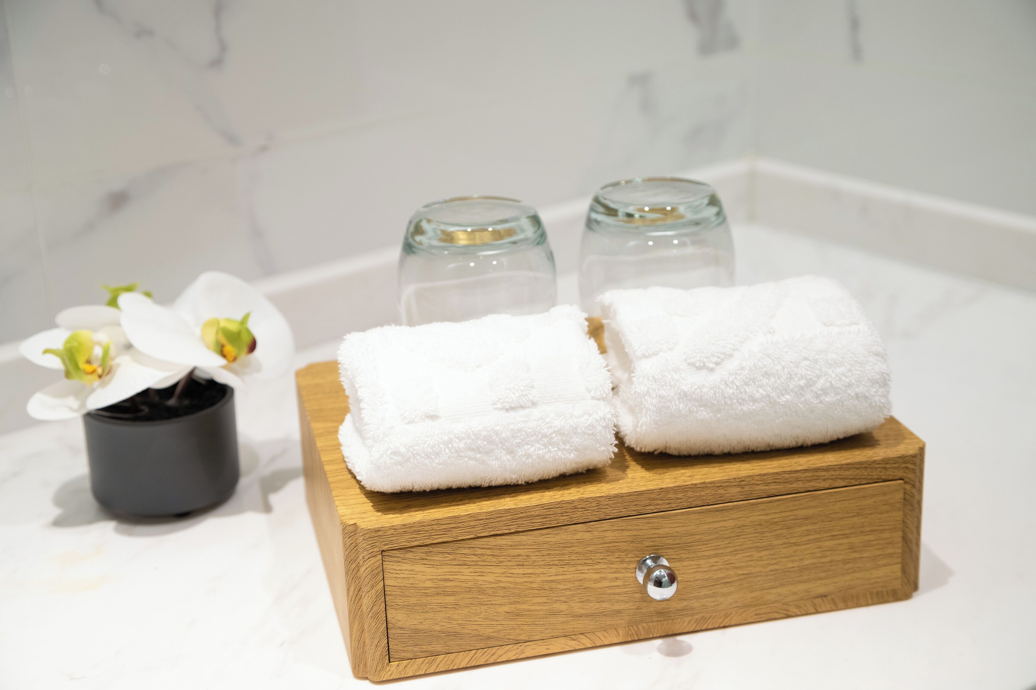 A wooden box with two folded white towels on a marble-tiled surface, a small potted orchid, and two clear glass bottles.
