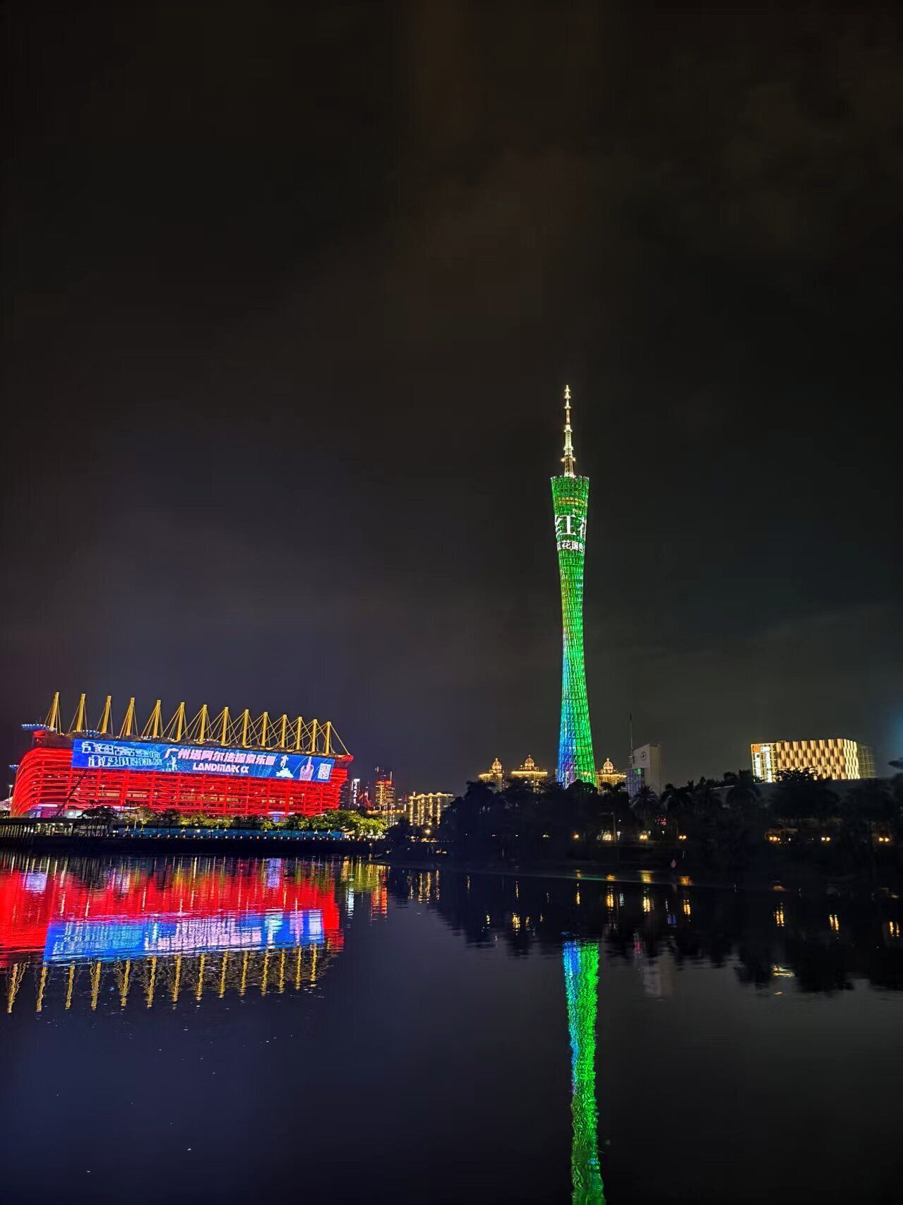Vista nocturna de un paisaje urbano con una torre destacada iluminada en verde y un edificio iluminado en rojo que se refleja en el agua.