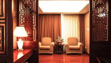 Habitación de hotel con dos sillones, una mesita con lámpara, un separador decorativo y un ventanal con cortinas.