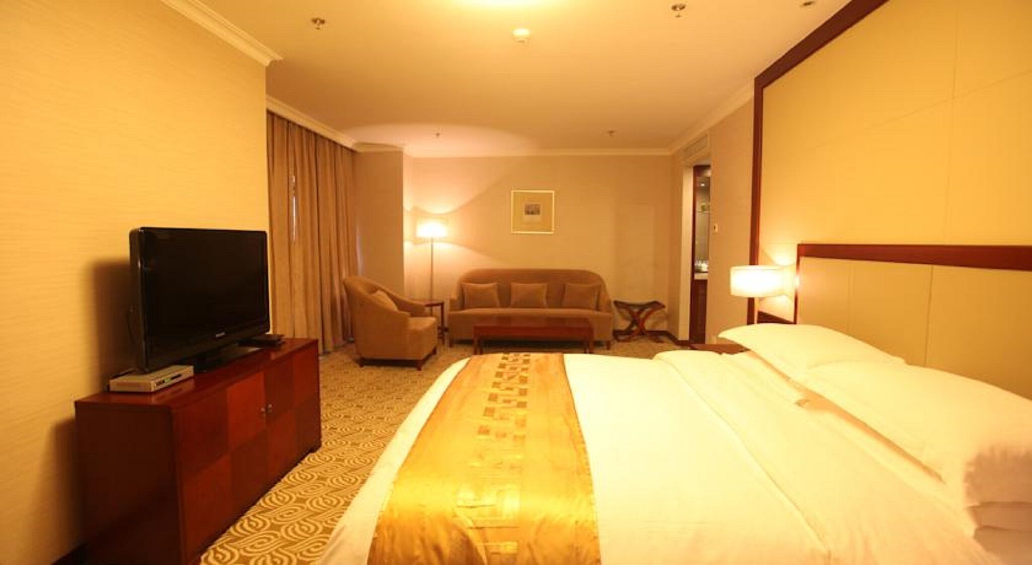 A hotel room with a bed, a television on a wooden cabinet, a sofa, a desk, and a lamp.